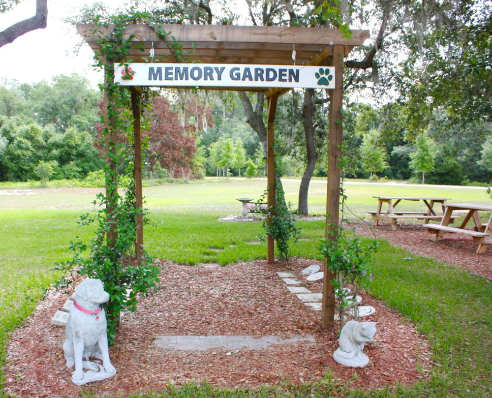 The Animal League Memory Garden arbor, bordered by memorial bricks and guarded by dog and cat statues. The area is shaded by trees with picnic benches underneath, and nearby a bird bath stands at the center of a flowerbed.