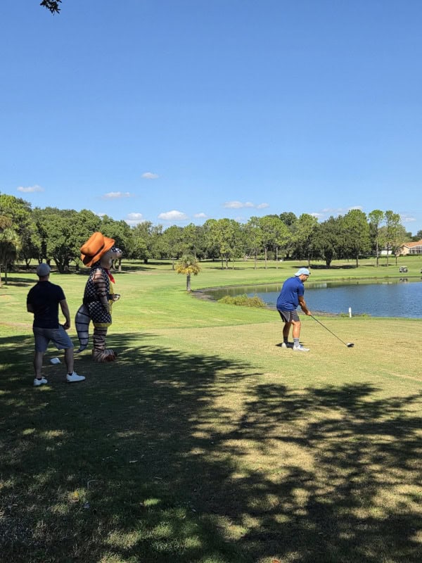 It's a beautifully, clear, sunny day as this golfer tees off. The grass looks lush and green beside a deep, blue lake, with a line of trees stretching into the distance.