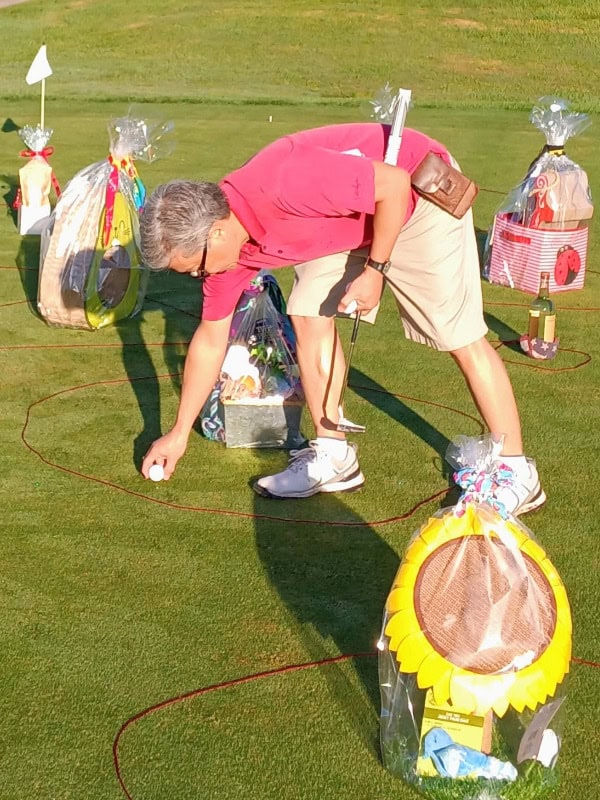 Gift baskets are placed on the green with red rope on the ground marking out an area around each prize. To win, players must hit a ball from the designated start point, into one of the prize areas. In this image a gentleman wearing a pink shirt is picking up his golf ball which he neatly hit into the area to win a selection of dog toys.