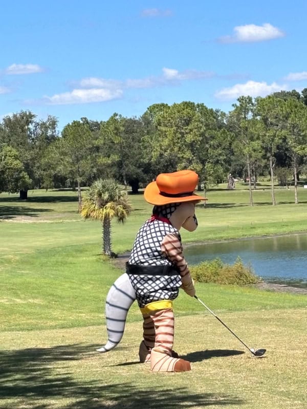 A person in a full-body cartoon armadillo mascot costume wearing an orange cowboy hat and a patterned vest, holding a golf club on a grassy golf course.