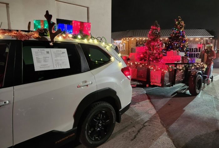 A white Subaru Estate, with lit-up presents, reindeer antlers, and more lights and tinsel, pulls our parade trailer through the streets.