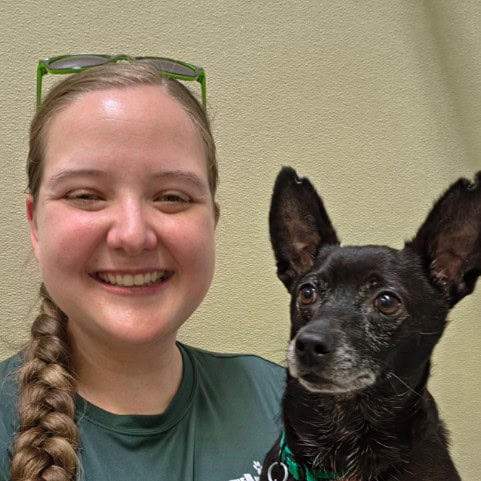 Shannon smiles broadly, wearing a green, Animal League T-shirt, glasses on her head, and plaited hair over one shoulder. She is cuddling Baloo, a black Chihuahua, who is looking interested in something beyond the camera.